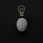 A gray brain model with a light bulb outline above it on a black background.
