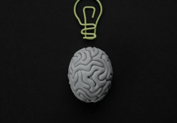 A gray brain model with a light bulb outline above it on a black background.