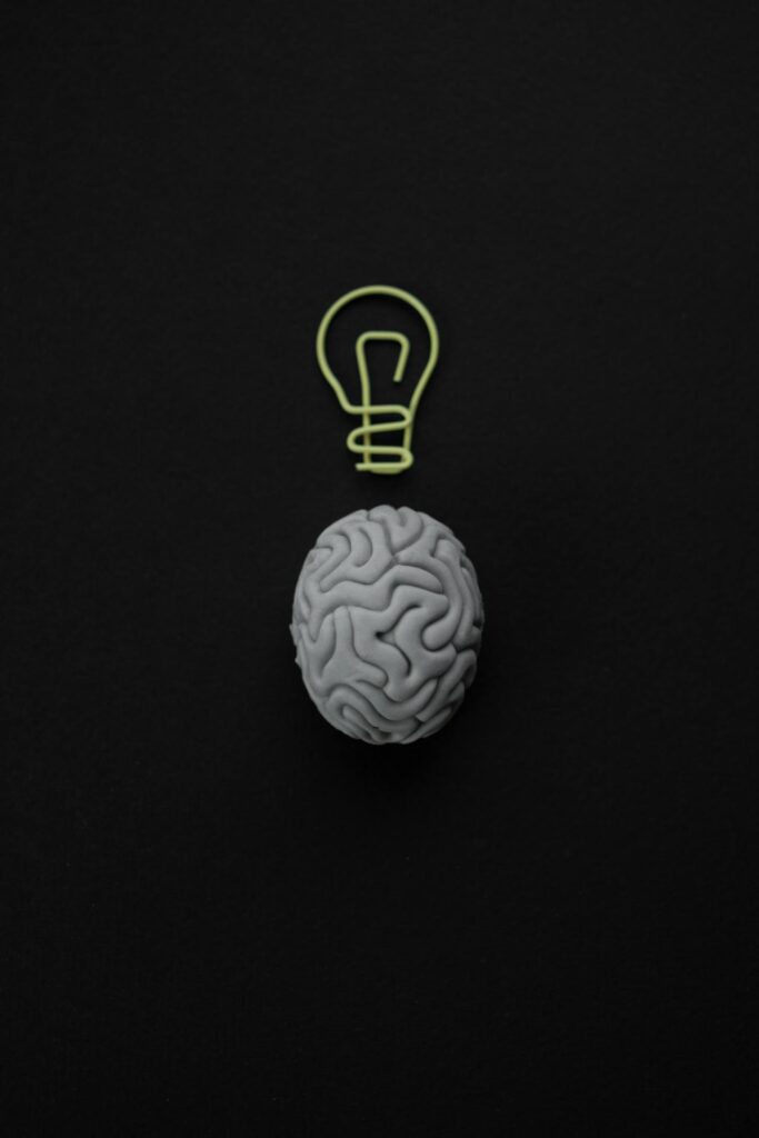 A gray brain model with a light bulb outline above it on a black background.